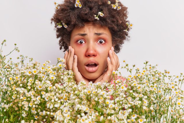 A person with flowers in their hair and a distressed facial expression stands behind a field of white daisies, holding their cheeks with both hands. A person with flowers in their hair and a distressed facial expression stands behind a field of white daisies, holding their cheeks with both hands.