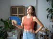 Why Is My AC Not Cooling? Common Causes and Solutionsf with a blue hand fan inside a room with potted plants and framed items in the background. She wears an orange sleeveless top and blue jeans.