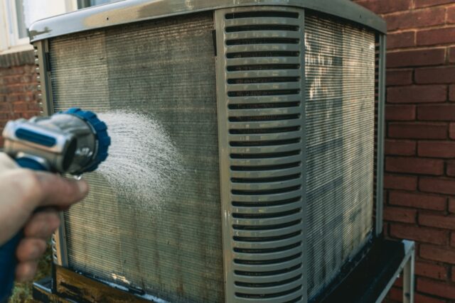Person cleaning an outdoor air conditioning unit using a hose. Clean Your Air Conditioner