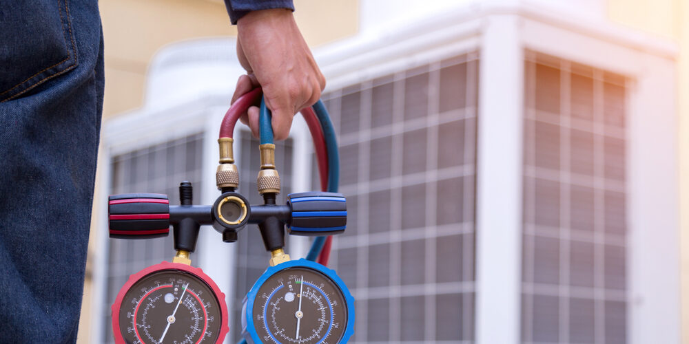 A person holds an HVAC manifold gauge set in front of large air conditioning units. cooling repair