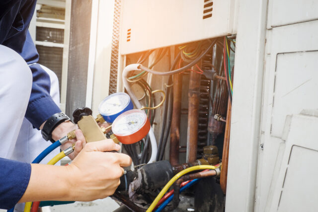 A technician is checking HVAC system gauges, holding red and blue testing tools connected to pipes and wires. air conditioning