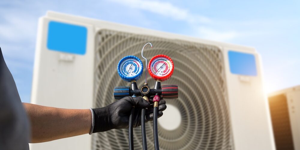 A gloved hand holds HVAC manifold gauges in front of a large air conditioning unit with blue sky in the background. Air Conditioner Maintenance