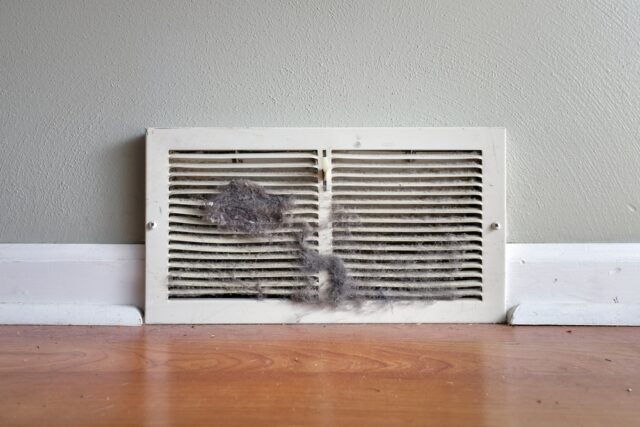 A white floor vent cover against a gray wall is clogged with dust and debris, standing above a wooden floor and white baseboard. dirty air duct air duct cleaning venice fl