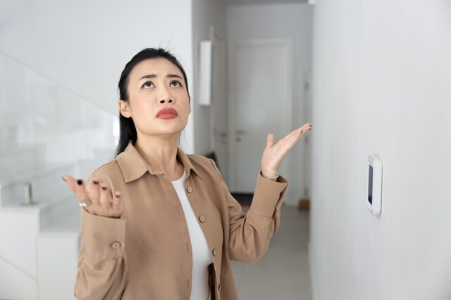 A woman stands indoors with her arms raised and looking up, appearing confused or frustrated, near a wall-mounted device. confused woman ac myths debunked