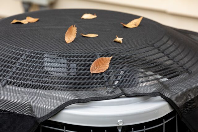 A close-up of an outdoor air conditioning unit with a mesh cover, scattered with several dry leaves on top. 7 Essential HVAC Prep Tips
