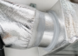Close-up of an insulated HVAC duct connection made of shiny, metallic material, sealed with tape and fitted to a rectangular air vent after air duct repair.