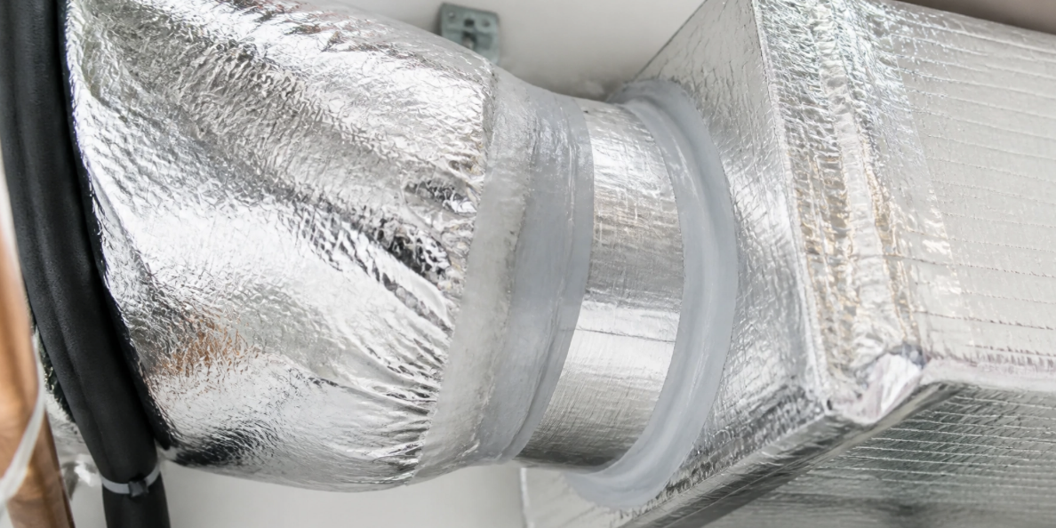 Close-up of an insulated HVAC duct connection made of shiny, metallic material, sealed with tape and fitted to a rectangular air vent after air duct repair.