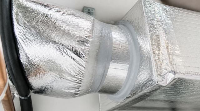 Close-up of an insulated HVAC duct connection made of shiny, metallic material, sealed with tape and fitted to a rectangular air vent after air duct repair.